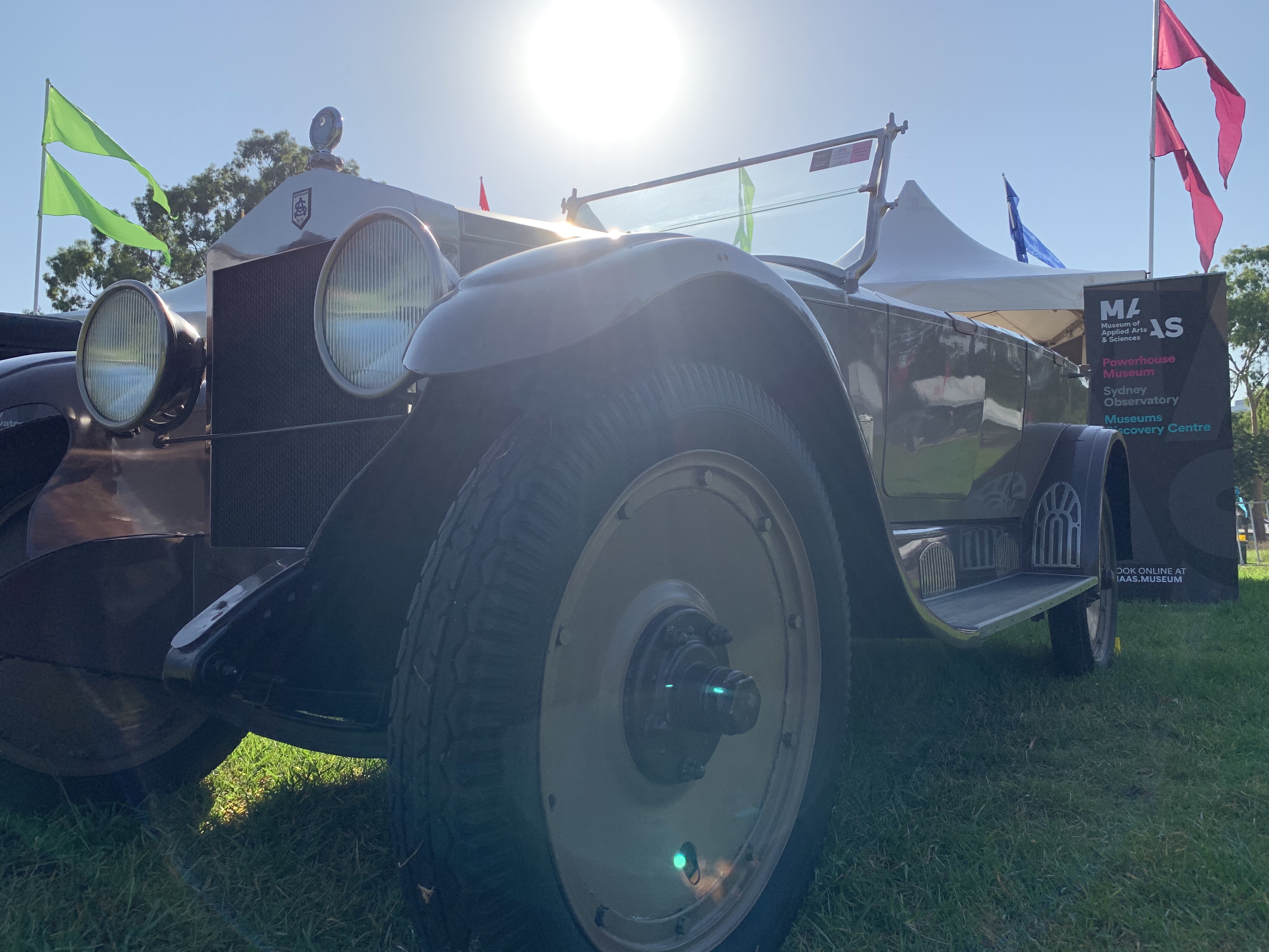 Low angle close shot of a dark and light brown classic car on grass. Front left wheel is the closest object in the photo. The sun is at the centre of the top section of the photo. 